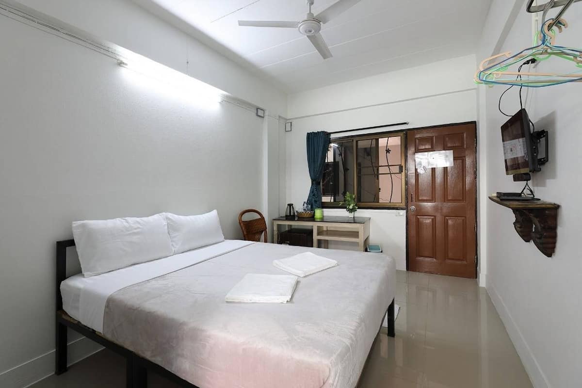Un dormitorio con cama, escritorio, silla, ventana con cortinas, puerta y televisor montado en la pared.