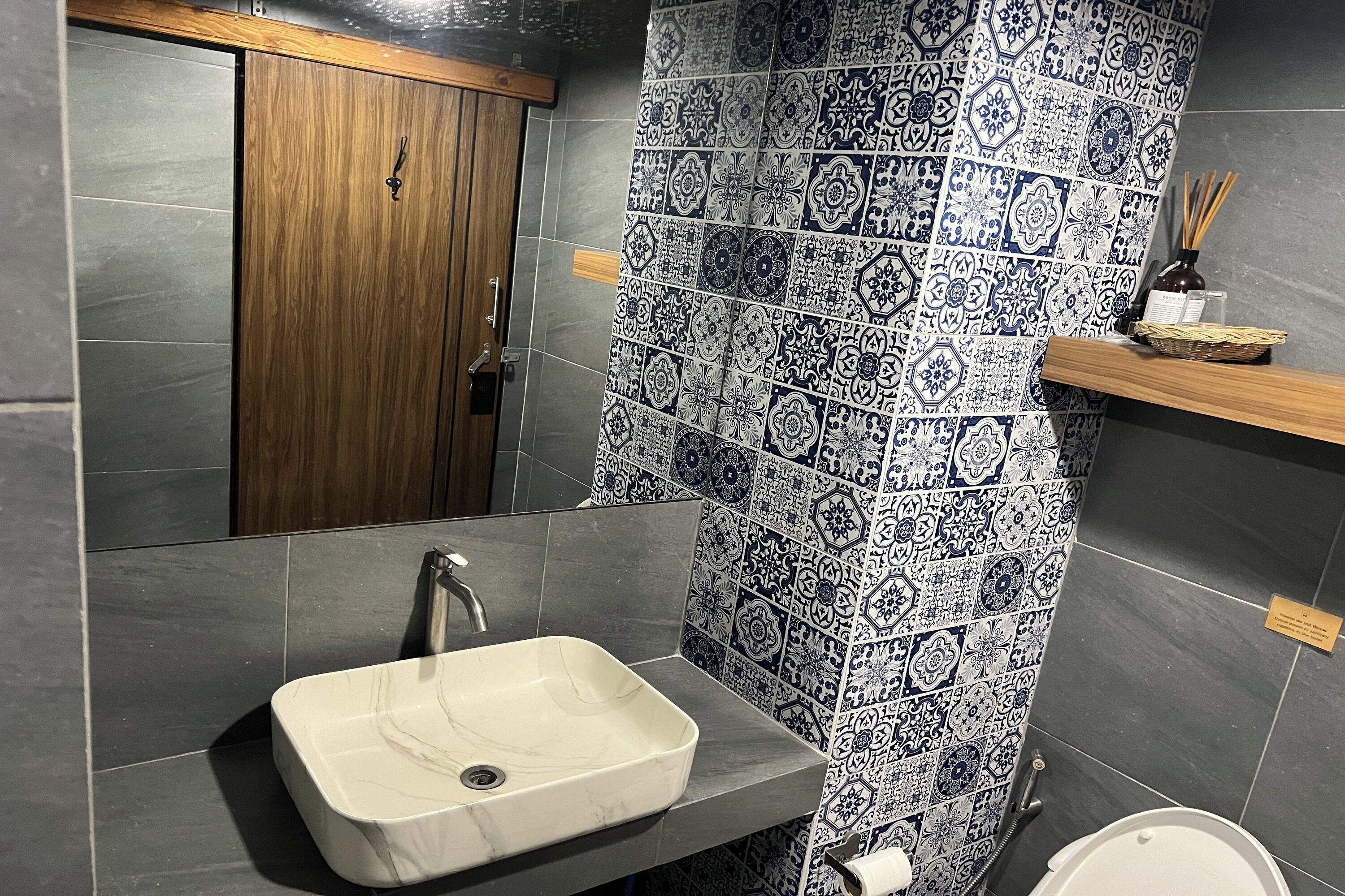 Une salle de bain avec un lavabo en marbre, des murs recouverts de carreaux foncés, une porte en bois et une étagère sur laquelle se trouvent un flacon et un diffuseur à bâtonnets.