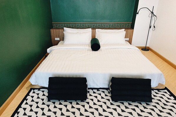 Un lit bien fait, avec du linge de lit blanc et deux oreillers noirs, un mur vert orné d’un motif décoratif et une lampe sur pied à droite.