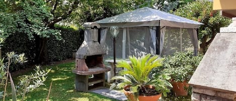 Un jardín con zona de barbacoa de piedra, un área de estar techada y diversas plantas en macetas.