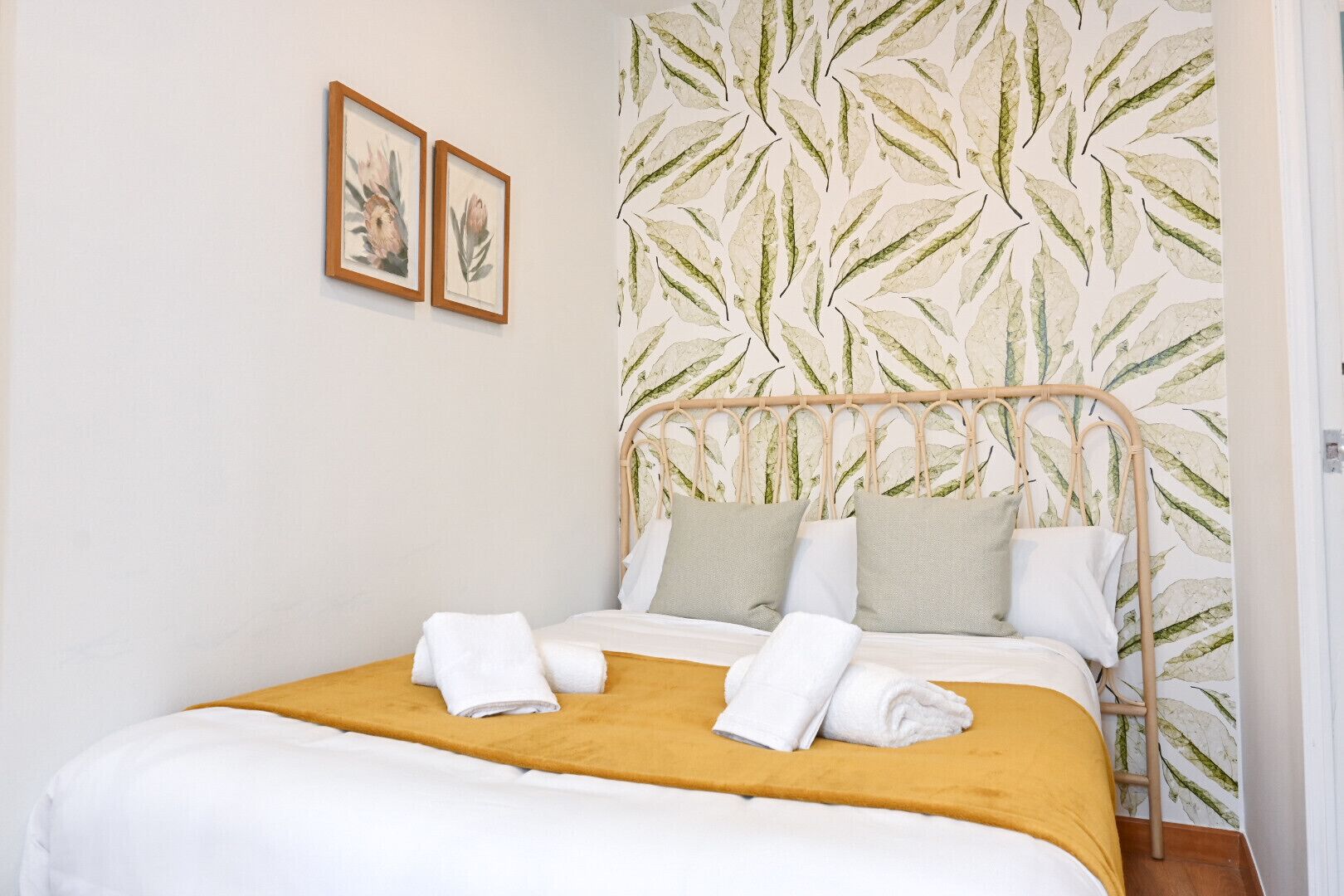 A neatly made bed with white bedding and a mustard-colored throw, a headboard with a leaf-patterned wallpaper, and two framed botanical prints on the wall.