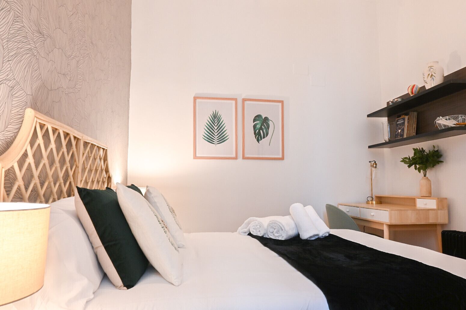 A neatly arranged hotel room with a bed, pillows, a bedside table with a lamp, a desk with a chair, and two framed leaf artworks on the wall.