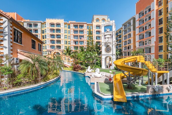 Relax by the resort pool surrounded by lush greenery and palm trees, with a central water slide and stunning views of the surrounding buildings.