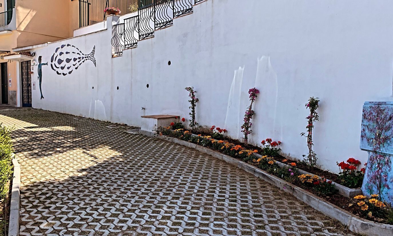 Un camino pavimentado con un diseño de baldosas, una pared blanca con un mural de peces y una fila de macizos de flores con diversas flores.