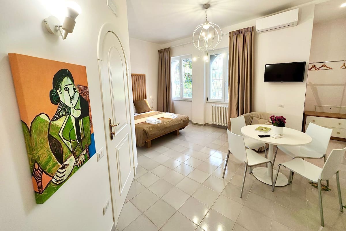A modern hotel room with a bed, a dining table with chairs, a TV, and a painting on the wall.