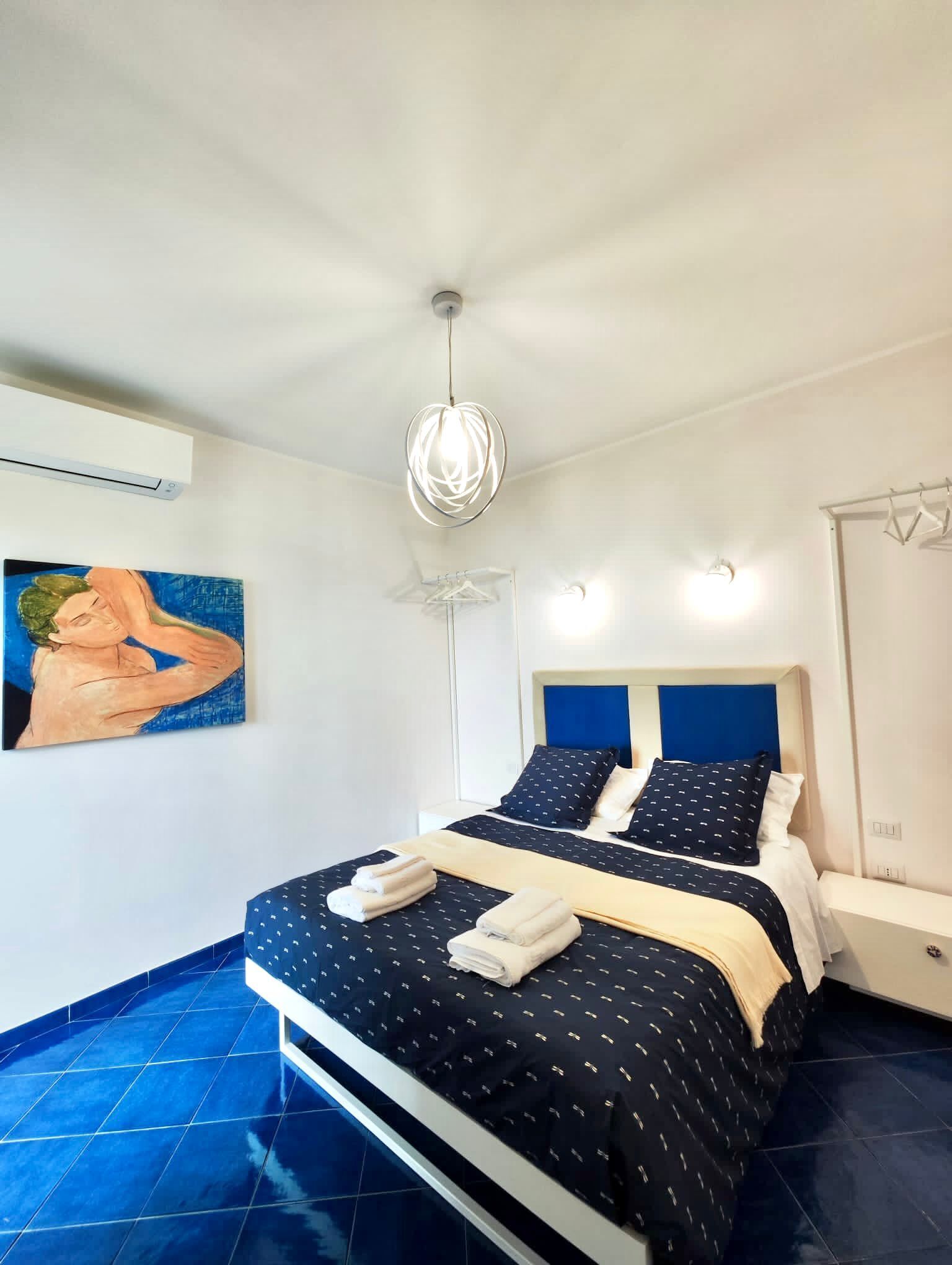 A hotel room with a bed, a painting on the wall, a ceiling fan, and a light fixture.