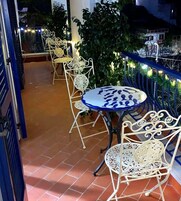 Una terraza con sillas de metal blanco y una mesa redonda, rodeada de plantas y luces colgantes.