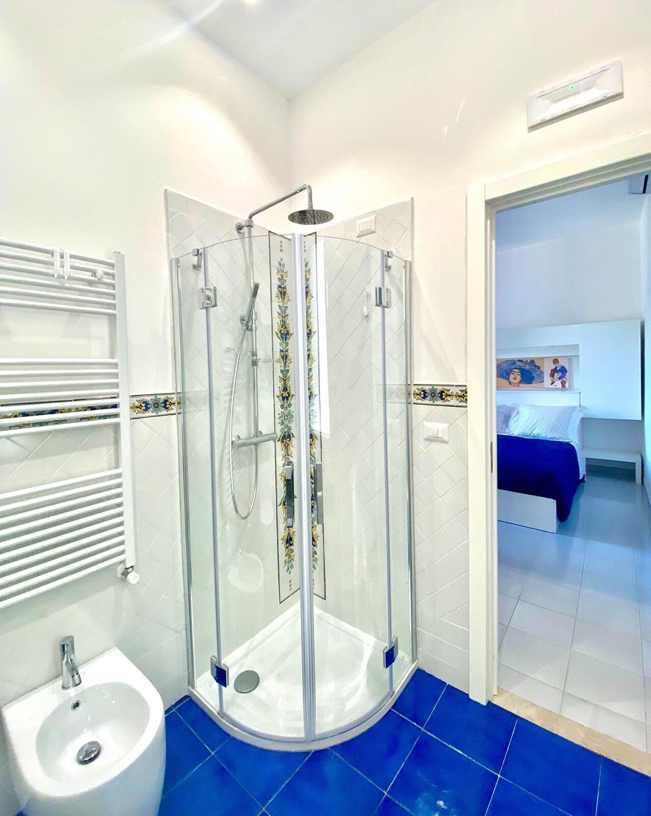 Un baño moderno con ducha de vidrio, lavamanos blanco y bidé. El piso tiene azulejos azules y un radiador blanco.