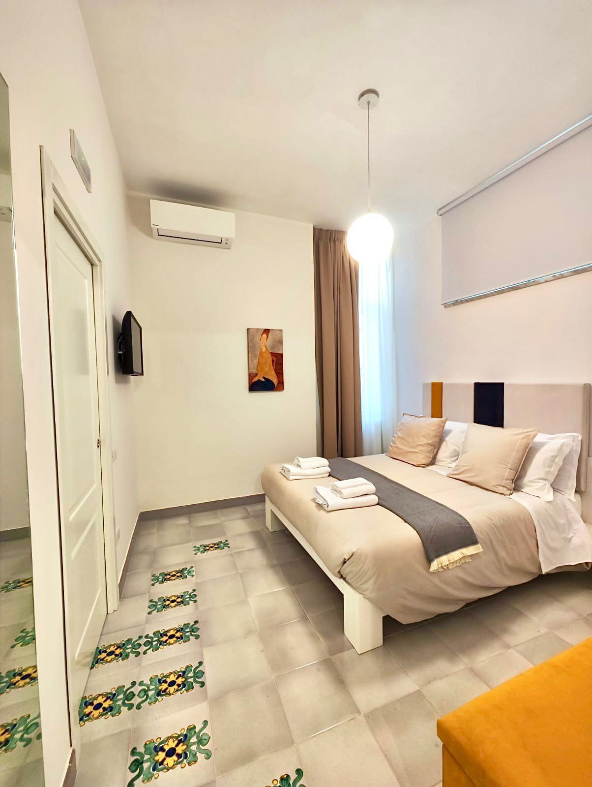 A hotel room with a bed, a bedside table, a flat-screen TV, a painting on the wall, and a window with curtains.