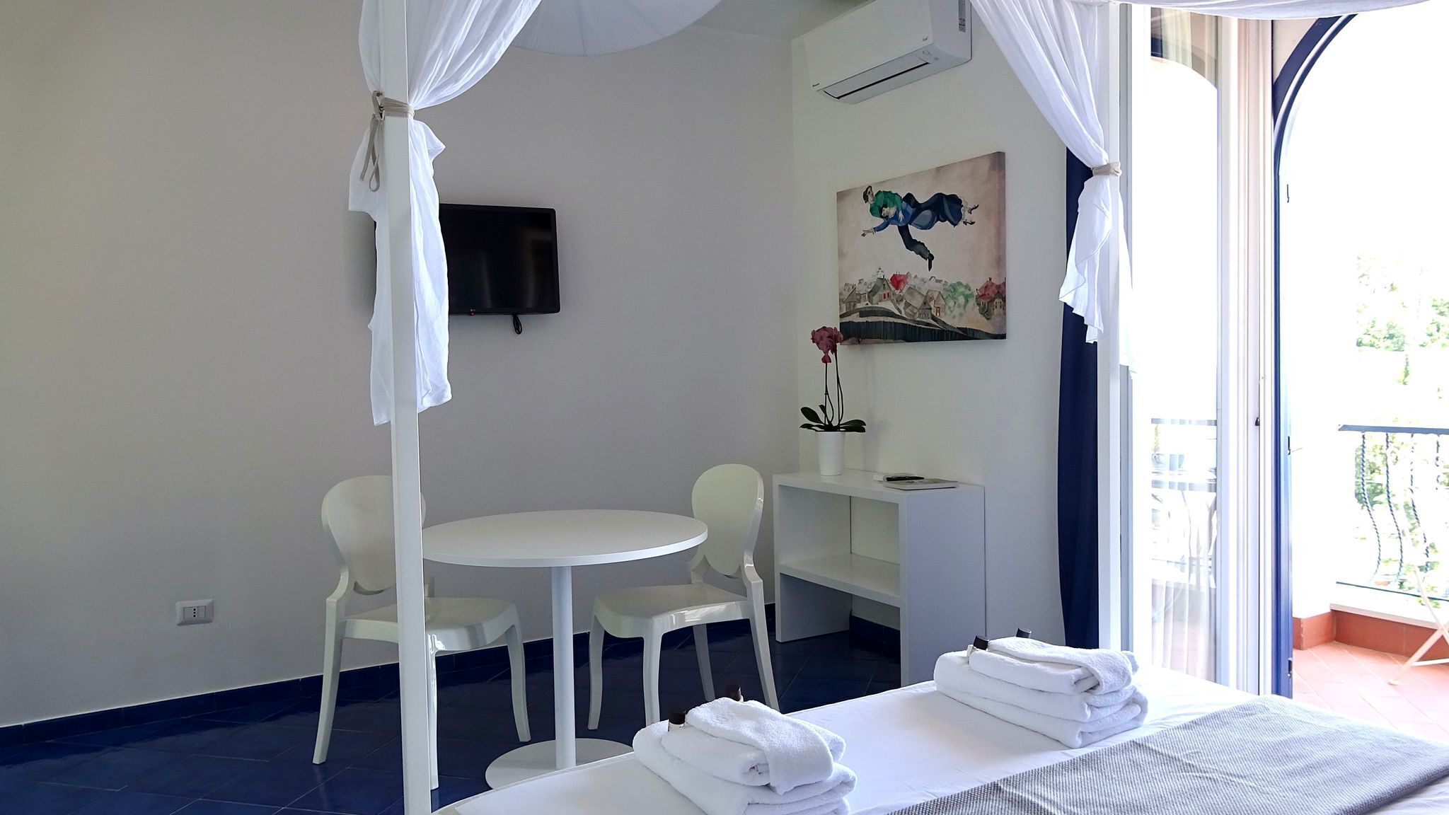 A hotel room with a bed, a small round table, two chairs, a TV, a painting, and a balcony with curtains.