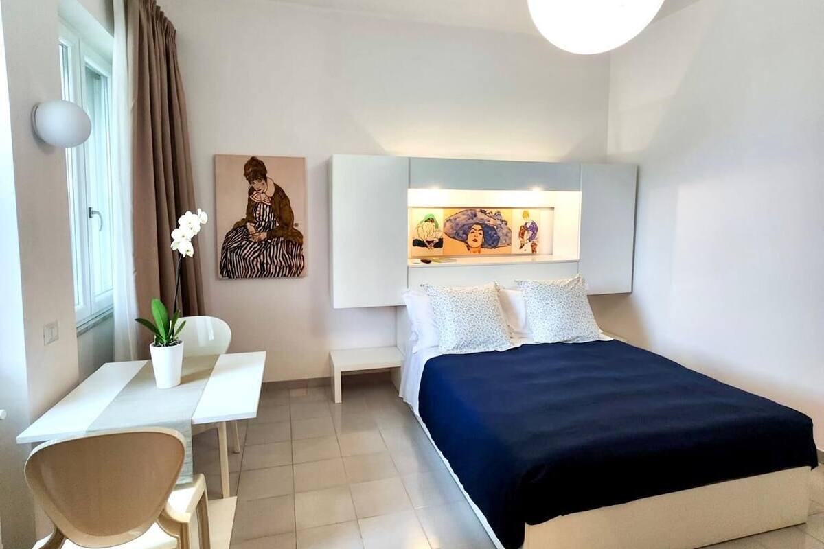 A modern hotel room with a bed, a desk, and a chair. There is a window with curtains, a painting on the wall, and a ceiling light.