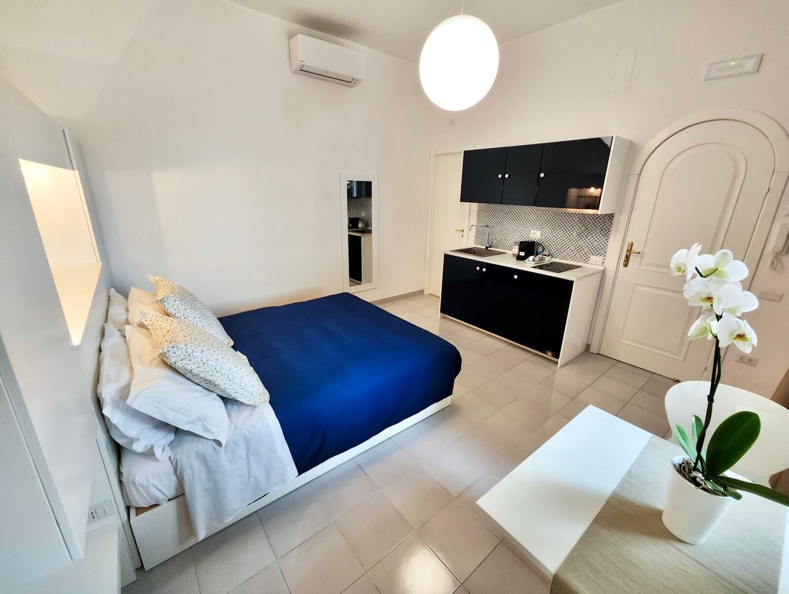 Ein gemütliches Schlafzimmer mit Bett, Nachttisch und einer Topforchidee. Angrenzend befindet sich eine Küchenzeile mit Herd, Spüle und schwarzen Schränken.
