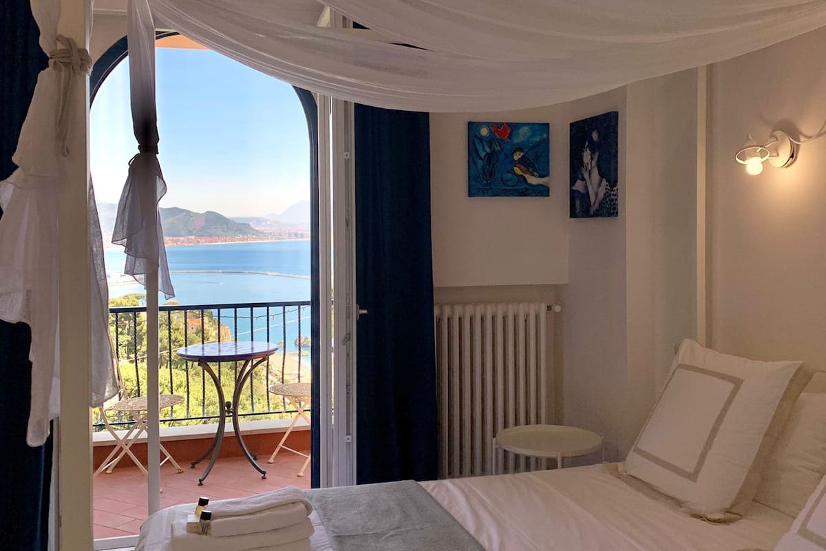 A bedroom with a bed, a small table, a chair, and a balcony with a view of the sea.