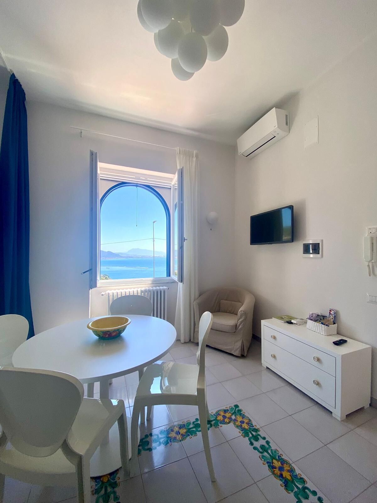 A modern, well-lit room with a round dining table, a beige armchair, a white dresser, and a TV mounted on the wall.