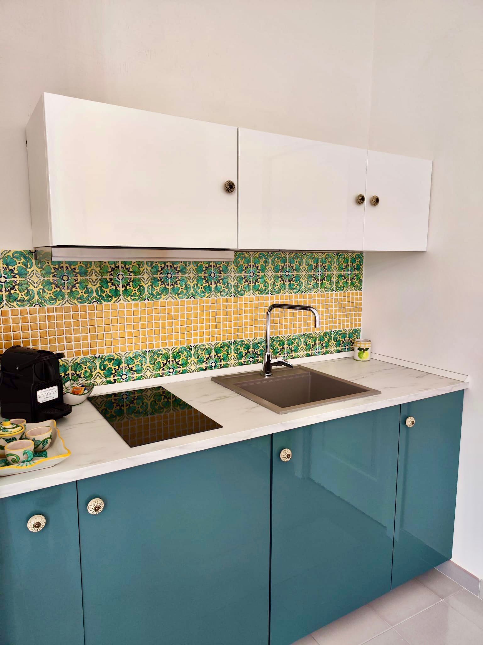 Une cuisine avec des meubles blancs, un évier et un crédence ornée de carreaux verts et jaunes.