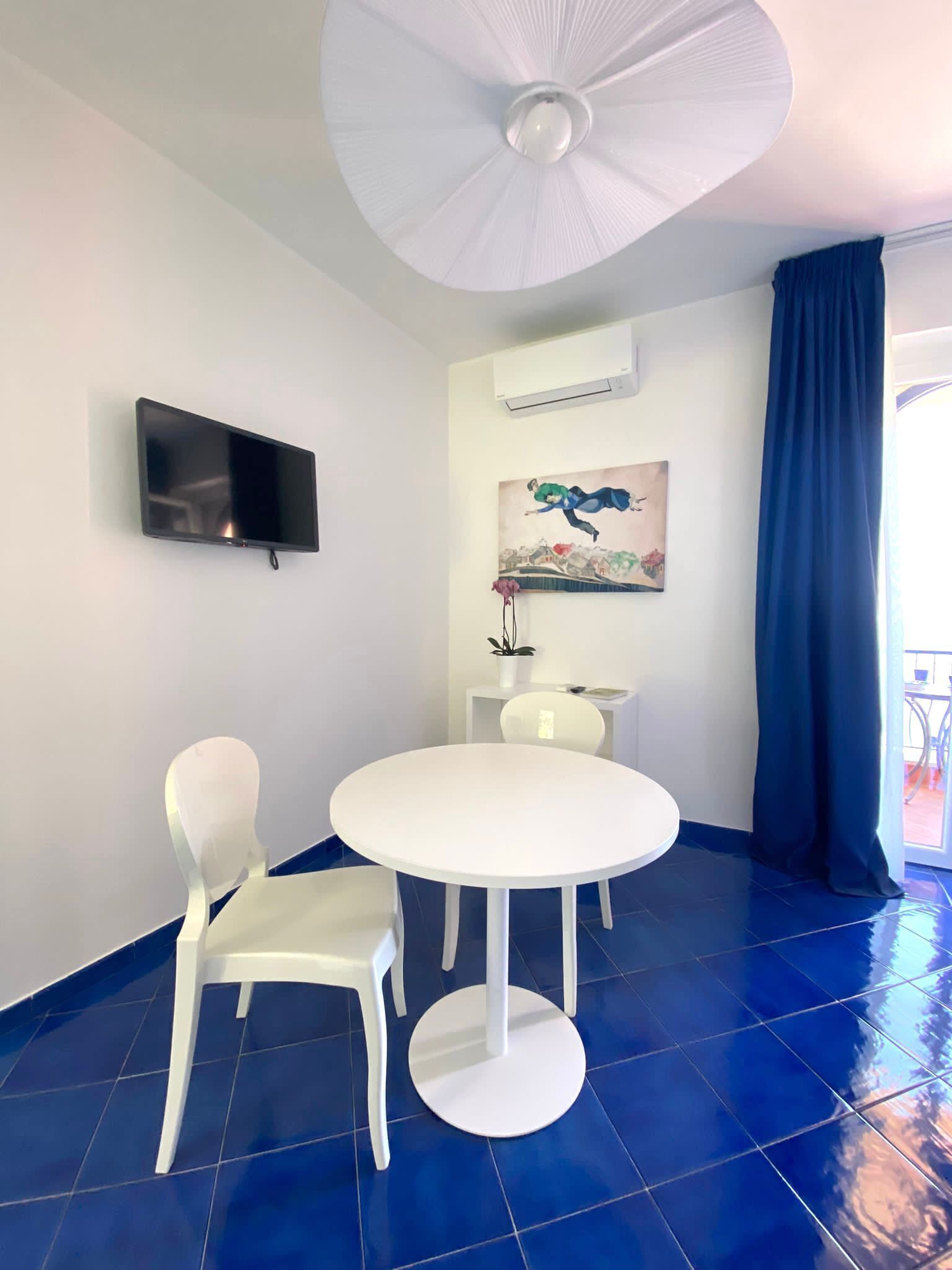 A room with a white table and chairs, a ceiling fan, a flat-screen TV mounted on the wall, and a painting above the table.