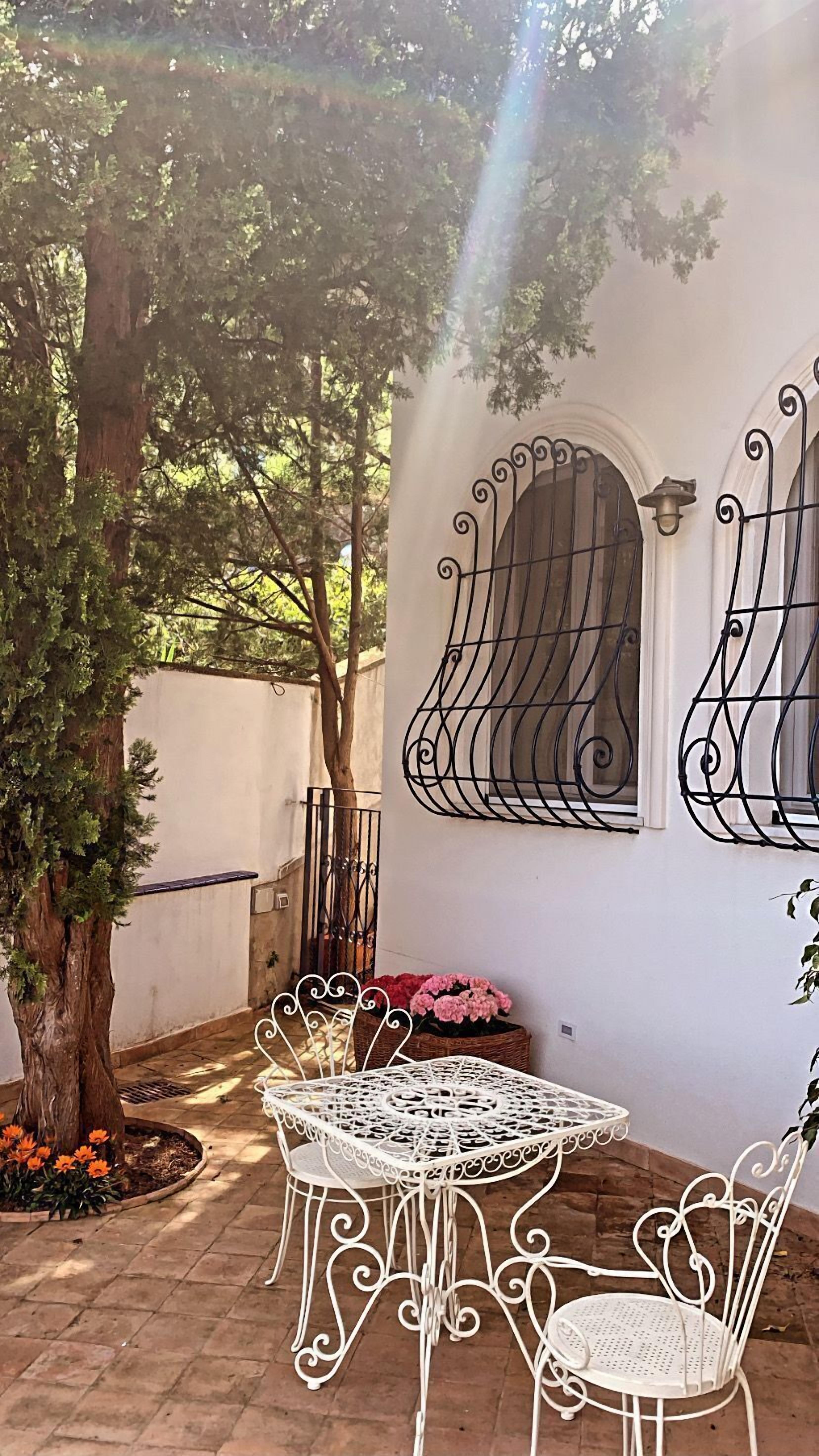 Een terras met een witte tafel en stoelen, een grote boom, en een muur met decoratieve metalen spijlen en een raam.