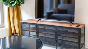 A modern living room with a flat-screen TV on a wooden stand, a glass coffee table, and a black metal console table with a plant.