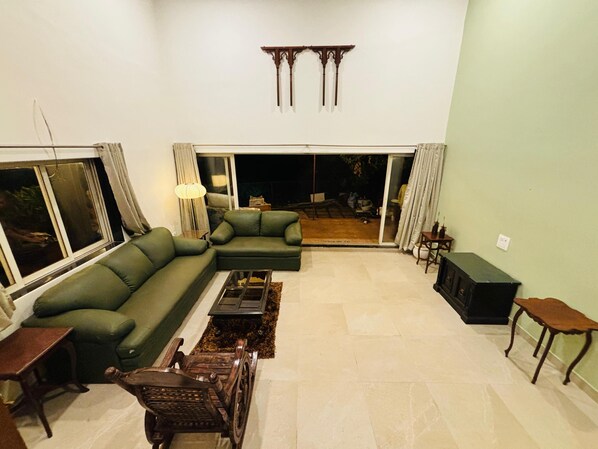 A living room with a sectional sofa, a coffee table, and a wooden chair. There is a sliding glass door leading to an outdoor area. - CooCasa (Mahabaleshwar)