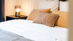 Una cama bien hecha con sábanas blancas y un edredón estampado, una lámpara de noche y un cabecero con un diseño oscuro y texturizado.