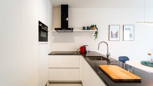 Una cocina moderna con armarios blancos, encimera negra, tabla de cortar de madera, hervidor rojo y dos cuadros en la pared.