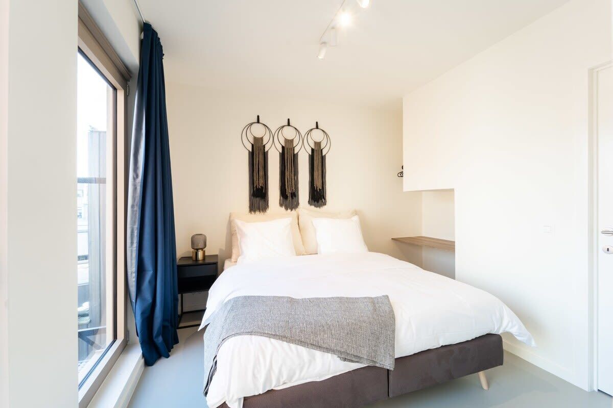 Un dormitorio moderno con una cama grande, una mesita de noche, una lámpara fijada a la pared y vistas al exterior a través de una ventana con cortinas.