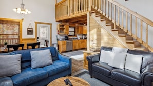 A spacious living area with a blue sofa, a black leather sofa, wooden flooring, a staircase, and a kitchen area. - 3 BR (11 Guests) near Parkway - WiFi, Hot Tub, Fire Pit, Game Room, Free Tickets (Sevierville)