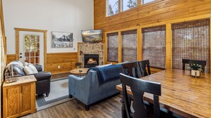 A cozy cabin interior with a fireplace, wooden walls, a dining table, and comfortable seating. - 3 BR (11 Guests) near Parkway - WiFi, Hot Tub, Fire Pit, Game Room, Free Tickets (Sevierville)