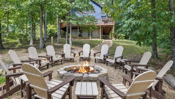 A circular fire pit surrounded by Adirondack chairs in a forested area with a wooden cabin in the background. - 3 BR (11 Guests) near Parkway - WiFi, Hot Tub, Fire Pit, Game Room, Free Tickets (Sevierville)