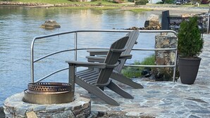 Un área de estar al aire libre con un hogar de piedra y sillas Adirondack junto a un lago.
