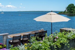 A table and chairs under an umbrella by a waterfront with a view of the sea and a lighthouse in the distance.