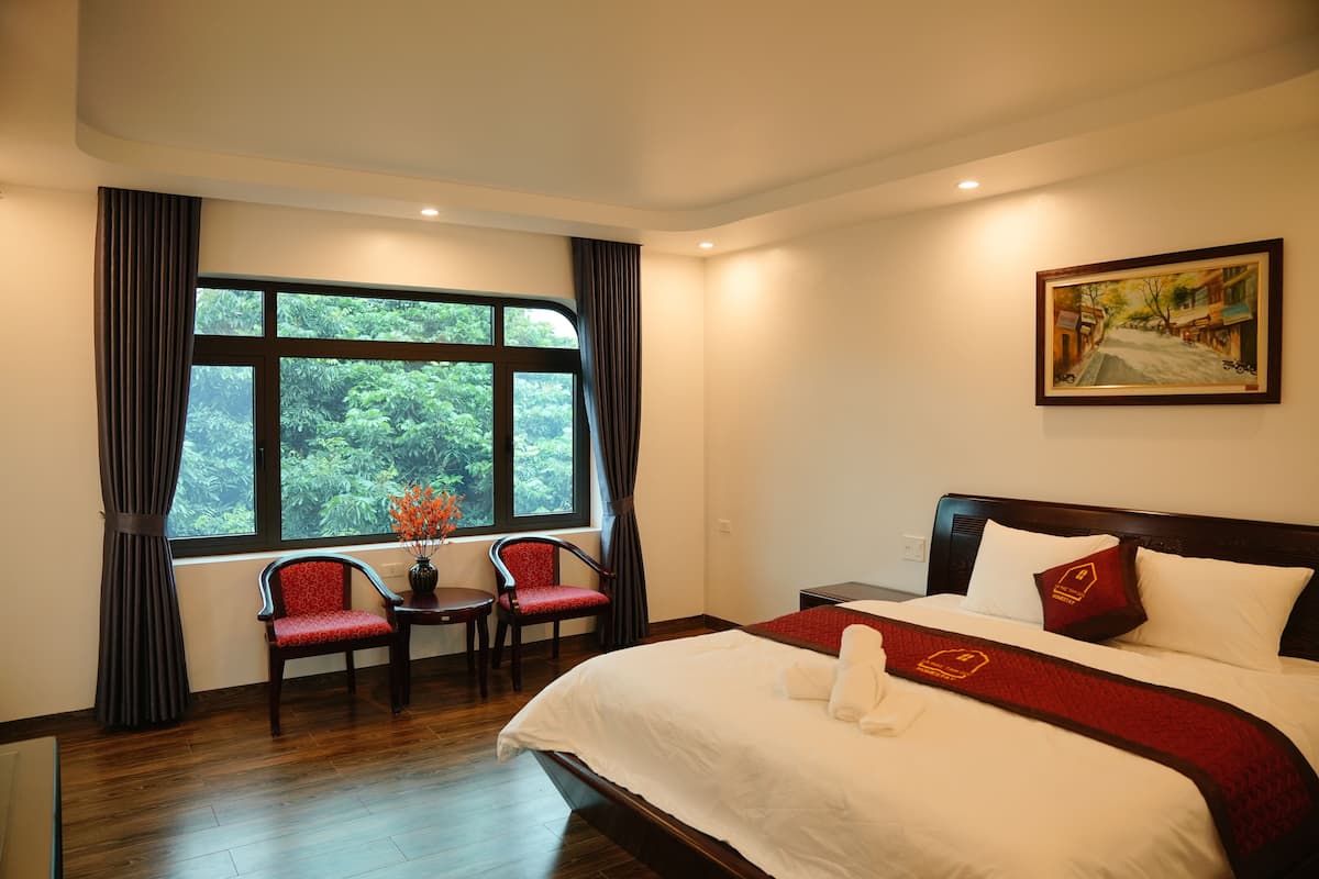 Quarto de hotel com uma cama grande, uma escrivaninha, duas cadeiras, uma janela com cortinas e um quadro na parede.