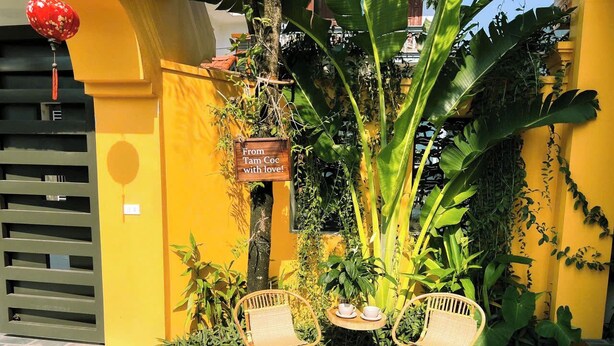 A yellow building with a sign reading "From Tam Coc with love!" and two wicker chairs with a small table and cups between them.