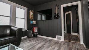Une pièce avec un canapé en cuir noir, une table basse en verre, une horloge, une guitare et un haut-parleur, le tout posé sur un plancher en bois.
