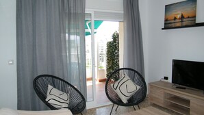 Un salon moderne avec un canapé, deux fauteuils noirs avec des coussins, un téléviseur posé sur un meuble en bois et un tableau représentant un coucher de soleil accroché au mur.