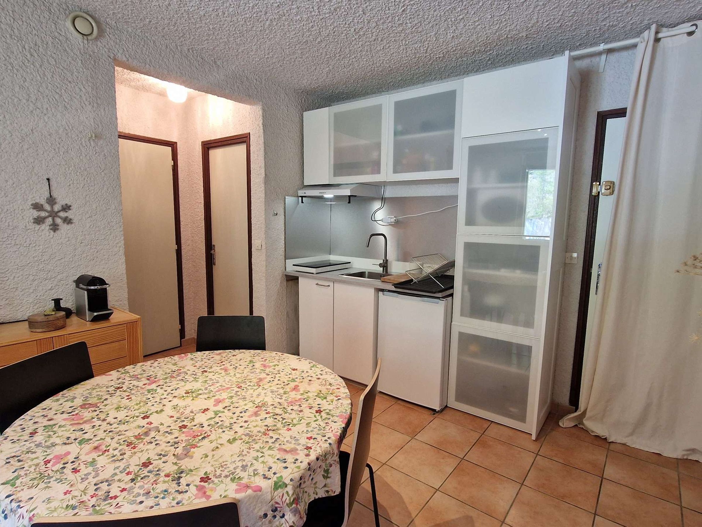 A compact kitchen with a dining table, chairs, a coffee machine, and a refrigerator.