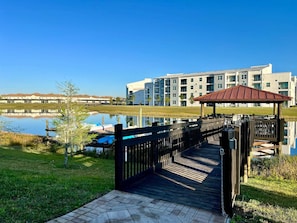 A wooden dock leading to a gazebo over a calm lake, with modern apartment buildings in the background. - Marvel | Mickey | Splash Pool | 4 Miles Disney 4876 (Kissimmee)