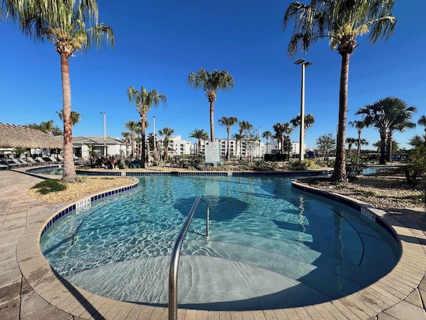 A clear swimming pool with a 2 ft 9 in depth, surrounded by palm trees and lounge chairs, with buildings in the background.