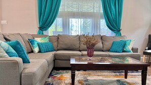 Ein Wohnzimmer mit einem grauen Ecksofa, türkisfarbenen Kissen, einer Glasvase mit getrockneten Blumen und einem farbenfrohen Teppich.
