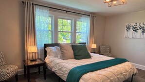 Ein Schlafzimmer mit einem Bett, zwei Nachttischen, einem Sessel und einem Fenster mit Vorhängen.