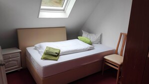 A neatly made bed with a white mattress, a green towel, and a white pillow. There is a wooden chair and a small bedside table with a lamp. - Room in Guest Room - Single Room With Shared Bathroom and Kitchen (Forbach)