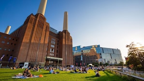 Un gran edificio industrial con dos chimeneas altas, una estructura moderna de vidrio y un área verde con gente sentada y caminando.