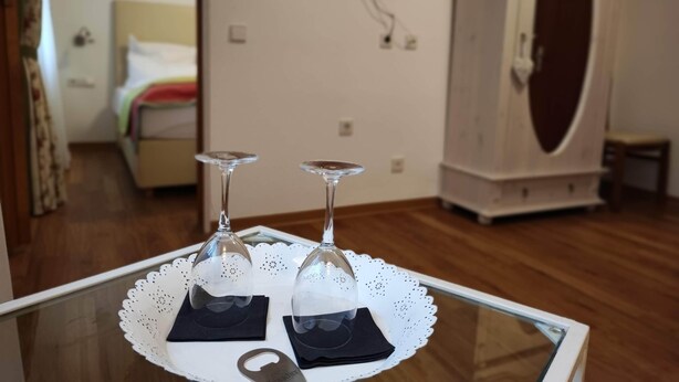 A room with a bed, a television mounted on the wall, a wardrobe, and a table with two wine glasses on a lace doily. - Room in Guest Room - Pension Forelle - Suite (Forbach)