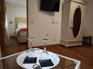 A room with a bed, a television mounted on the wall, a wardrobe, and a table with two wine glasses on a lace doily. - Room in Guest Room - Pension Forelle - Suite (Forbach)