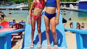 Dos personas de pie sobre una silla azul, con un letrero de playa al fondo.