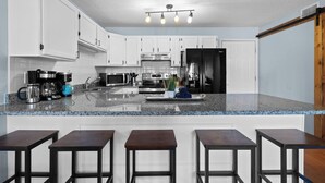 Una cocina moderna con armarios blancos, encimera de granito, cafetera, microondas, horno y refrigerador negro. Hay cuatro taburetes en la encimera.