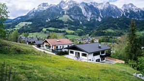 Une maison moderne dotée d’un balcon, entourée de champs verdoyants et avec des montagnes en toile de fond.