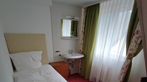 A hotel room with a bed, a bedside table, a mirror, a sink, and a window with curtains. - Room in Guest Room - Single Room With Shared Bathroom and Kitchen (Forbach)