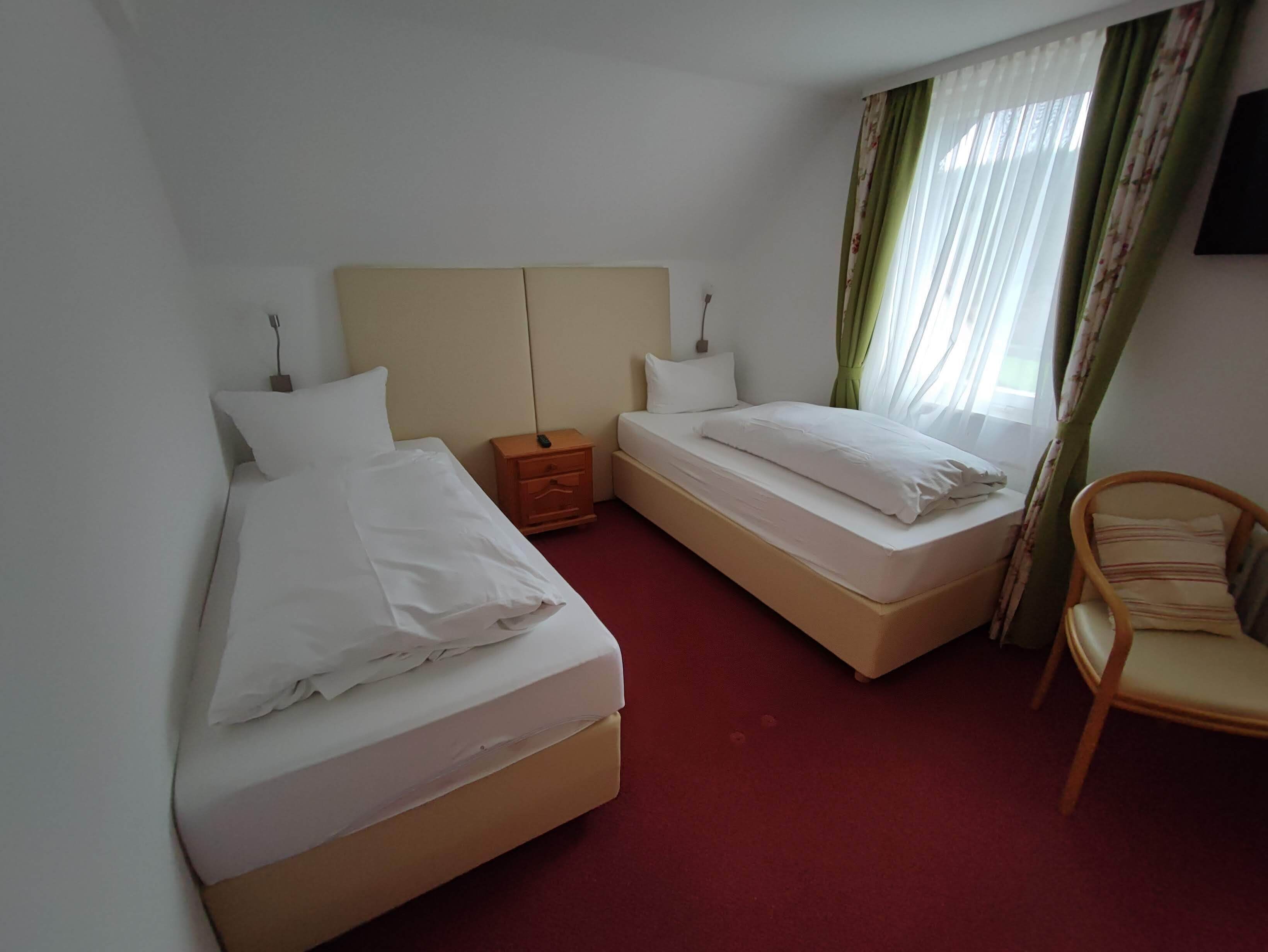 Ein Hotelzimmer mit zwei Einzelbetten, einem Holfnachtkästchen, einem Sessel und einem an der Wand befestigten Fernseher.