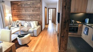 A modern living room with a wooden accent wall, a sofa, a coffee table, and a kitchen area with wooden cabinets and appliances. - Steinberghaus Top 3 (Rain)